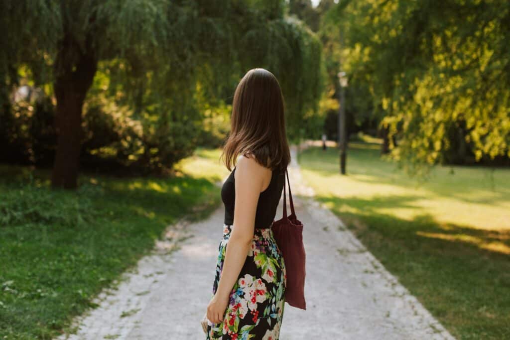 Woman with long hair walking on a park trail with trees and greenery.