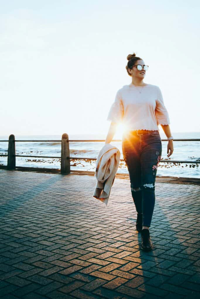 Stylish woman walking along the seaside promenade at sunset with sunglasses and casual outfit.
