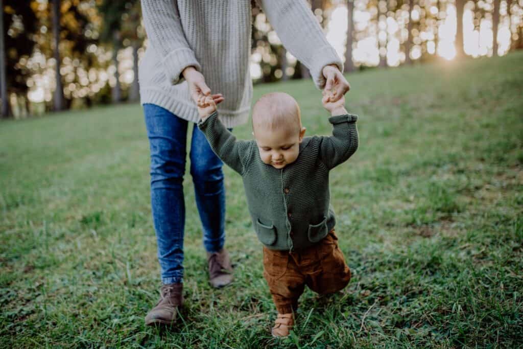 Child learning to walk with parent support outdoors in park.
