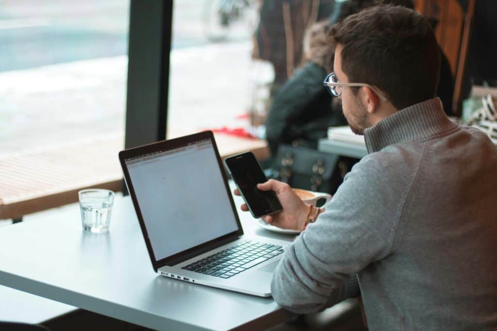 Man working on laptop and smartphone at cafe with large window view.