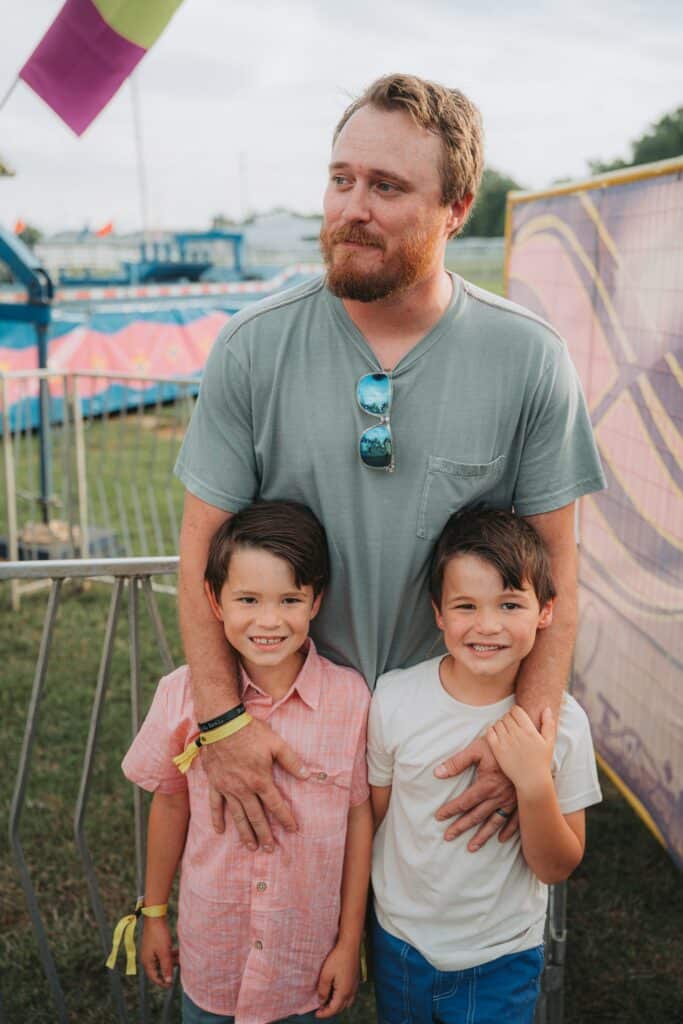 Father with two children at fair, smiling and enjoying outdoor event.