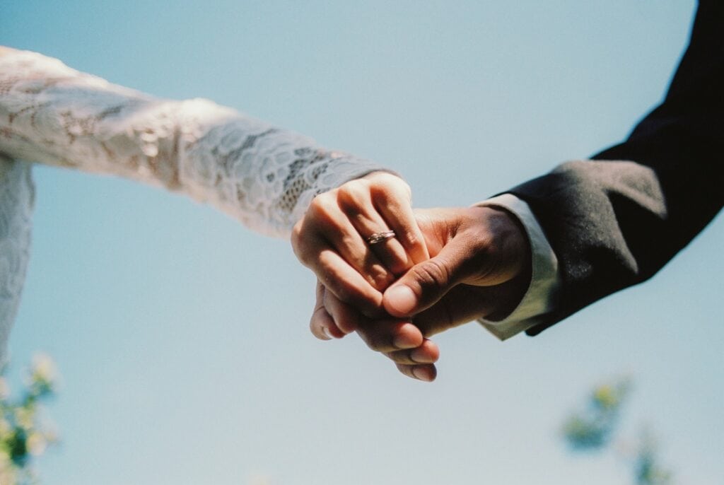 Close-up of a couple's hands in a marriage agreement, wedding dress and suit visible.