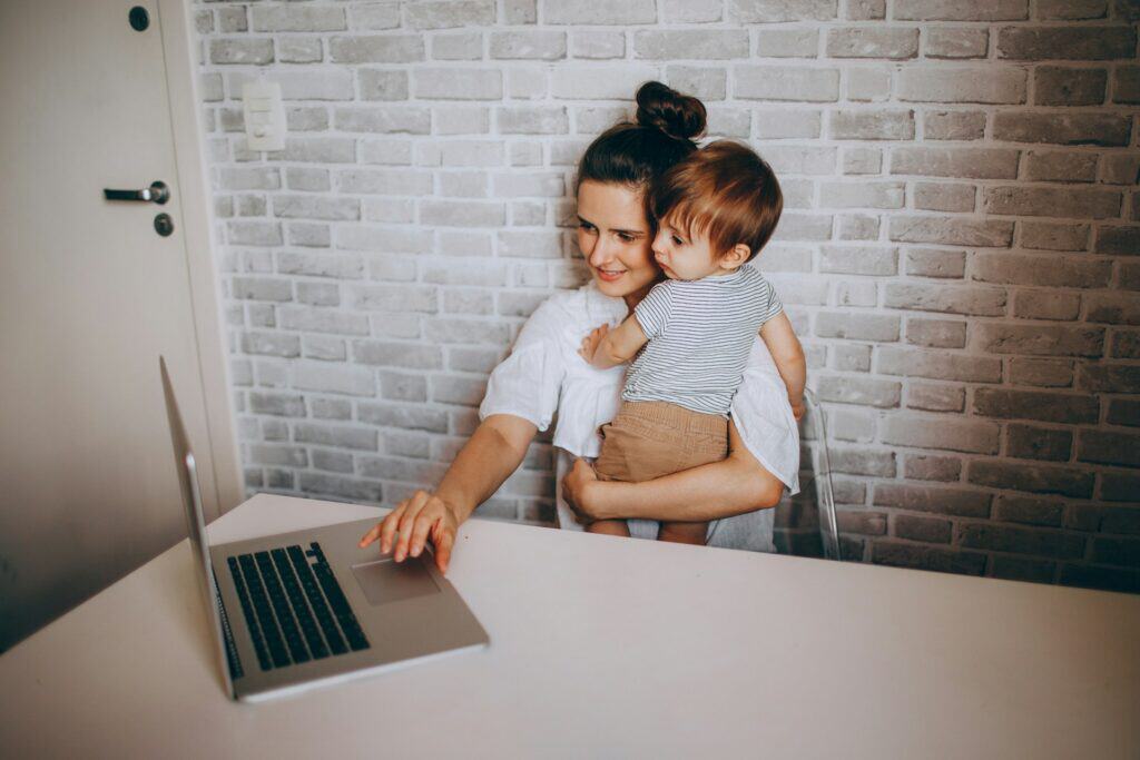 Mother working on laptop with young child in her arms.